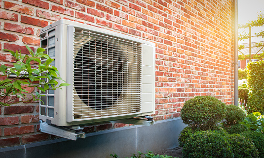 Side view of outdoor energy unit hanging on brick wall of house on a sunny day. Air to air heat pump for cooling or heating the home. Outdoor unit powered by renewable energy. Air conditioner and air heat pump.