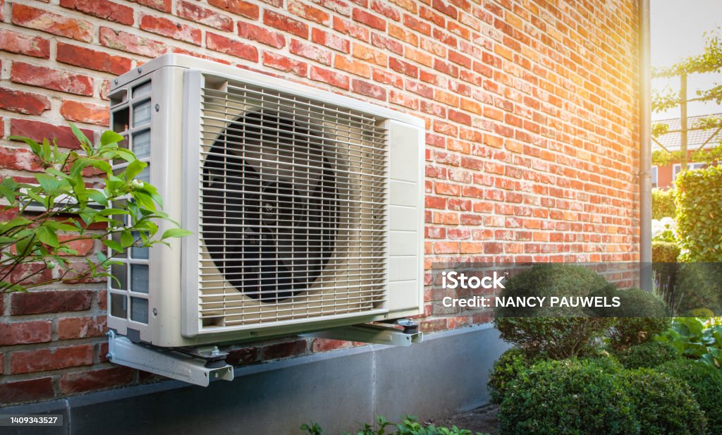 Air conditioning heat pump outdoor unit against brick wall. Side view of outdoor energy unit hanging on brick wall of house on a sunny day. Air to air heat pump for cooling or heating the home. Outdoor unit powered by renewable energy. Air conditioner and air heat pump. Heat Pump Stock Photo