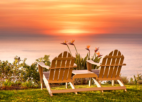 Two Adirondack beach chairs overlooking an orange sunset below the ocean horizon.