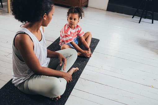 Latin mother and her little daughter doing yoga on a mat at home together.