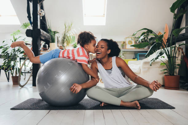 Mother And Daughter Exercising Latin woman sitting in a lotus pose on a mat, her little daughter on a pilates ball next to her, kissing her. mindfulness children stock pictures, royalty-free photos & images
