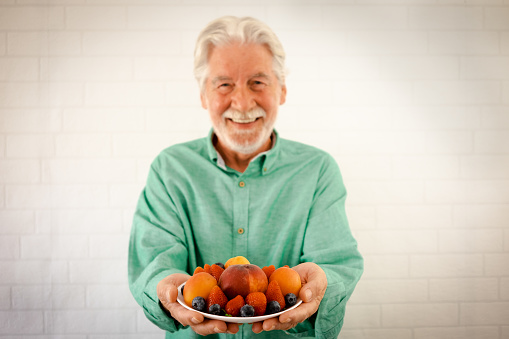 Defocused cheerful senior man holding a dish of fresh summer red fruits, elderly white bearded grandfather enjoying healthy eating