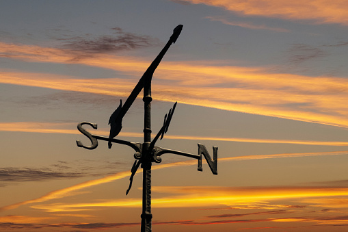 A weather vane points to the west with a sunset sky in the background in a composite image.