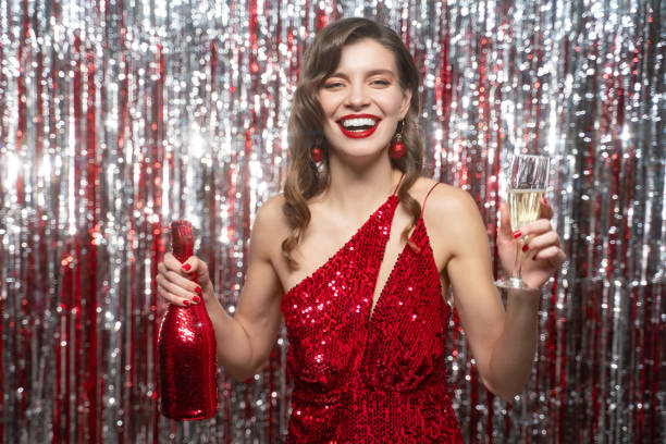 Happy woman celebrating christmas dancing with champagne on tinsel background in red dress Happy party woman celebrating new year dancing with glass and bottle of champagne on tinsel background, wearing red dress, lipstick and manicure woman in red dress stock pictures, royalty-free photos & images