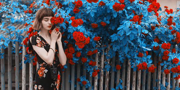 Redhead model on background of roses bush. Fabulous asian girl with red lips in dress on awesome summer background. Rose garden. Woman portrait. Valentines Day card. Spring flowers. Bob hairstyle
