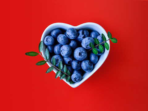 Creative summer layout of blueberries and natural green leaves in white heart shaped bowl on vibrant red background. Original blueberry decoration. Minimal summer concept. Welcome summer. Copy space.