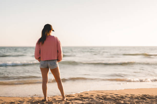 femme en sweat-shirt et casquette regarde le coucher de soleil sur la plage - sweat women wet shirt photos et images de collection