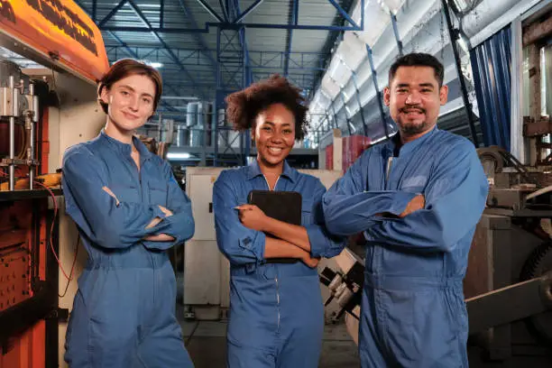 Cheerful multiracial industry workers in safety uniforms line up and arms crossed together after work success and express smiles and happiness in mechanical factory. Professional engineer occupation.