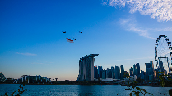 Singapore - July 16 2022: Chinook helicopters flying the Singapore National flag across the city's skyline during the National Day Parade rehearsal in the evening. Singapore state flag flying past the Singapore Flyer, Marina Bay Sands and Gardens by the Bay.