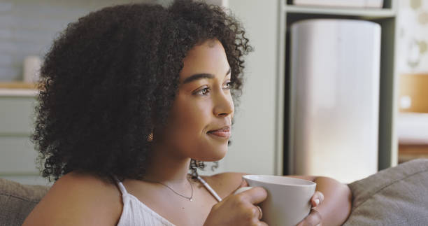 Beautiful black woman with an afro drinking a cup of coffee in the morning at home. Happy attractive female thinking while having tea on a couch in her house. A young lady relaxing on a sofa stock photo