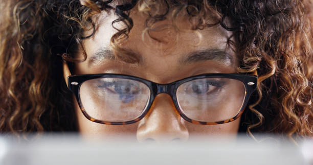 Closeup of a focused business woman with eyeglasses looking at laptop screen. Young worker with protective reading glasses checking emails or studying online. Details of face with trendy specs frame stock photo