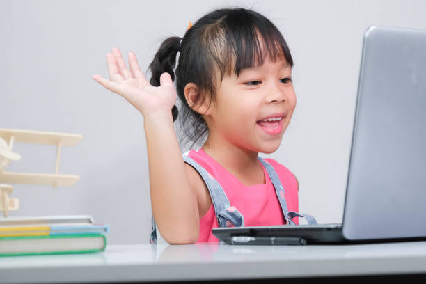 istock Smiling little Asian girl studying online having video call distant class with teacher using laptop. Happy little girl wave greeting with tutor while studying online with laptop at home. homeschooling 1409421084