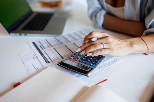 Close-up shot of unrecognizable young woman sitting at the table and using calculator while paying bills at home