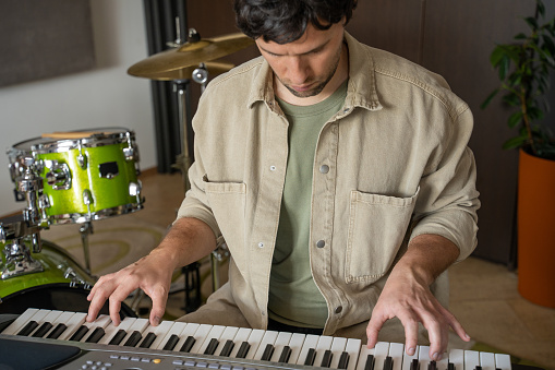 Man enthusiastically plays synthesizer in specifically equipped music studio. Guy in casual clothes and cap sits against drum-kits and cymbals