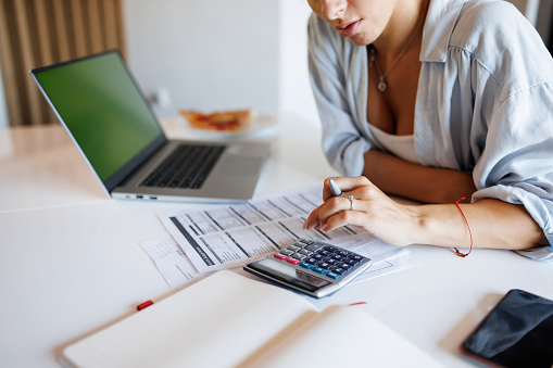 Close-up shot of beautiful concentrated young woman sitting at the table and using calculator while paying bills at home