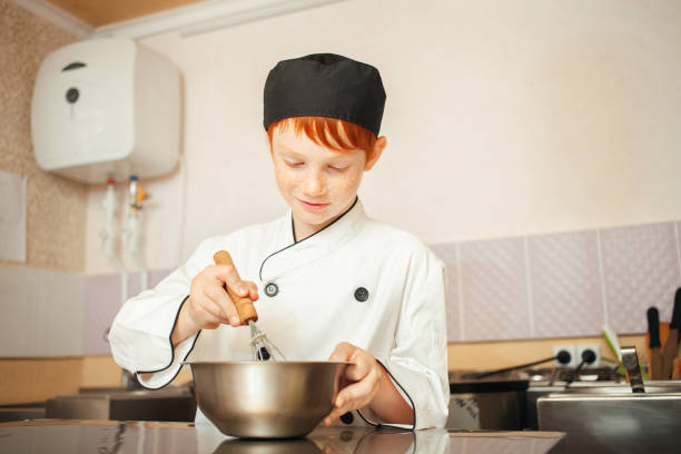 istock red-haired boy in chef's costume cooks pancakes in kitchen, kneads dough, assistant makes breakfast. Beat dough with whisk 1409424197