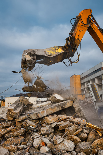 Contemporary excavator with hydraulic press breaks armored concrete leftovers over garbage against cloudy sky at demolition site