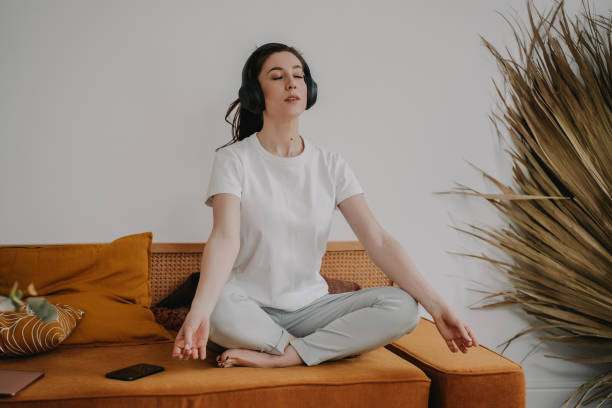 Indoor shoot of meditating beautiful woman sitting on orange sofa at home legs crossed at home with headphones. Girl relaxing after studies, peacefully sitting in a yoga posture. Calm housewife sesión interior de una hermosa mujer meditando sentada en el sofá naranja de casa con las piernas cruzadas en casa con auriculares. chica relajándose después de los estudios, sentada pacíficamente en una postura de yoga. ama de casa tranquila