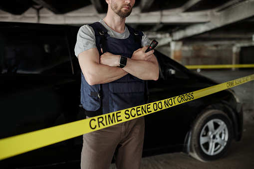 Cropped shot of crime scene investigator in bulletproof vest crossing arms by chest while standing behind yellow tape against car