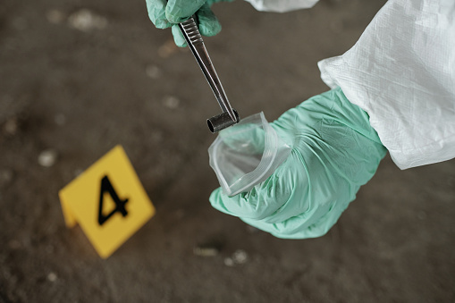 Gloved hands of criminological expert with metallic pincers putting empty cartridge case into plastic packet during inspection of evidences