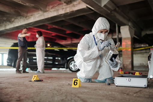 Young criminological expert in coveralls inspecting empty bottle on crime scene while squatting in front of open briefcase with working supplies