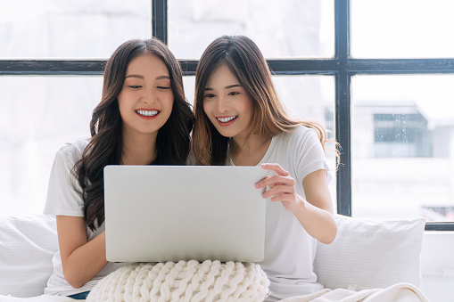Cheerful asian LGBTQ lesbian woman using laptop online sitting together with pillows white bed at apartment. Happy two young girl couple leisure with notebook working online. Technology communication