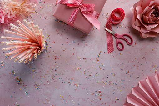 Directly above shot of a pink themed background with colorful sprinkles all around, a birthday gift, ribbon, drinking straws and paper decorations, arranged to have a copy space at the bottom of the shot.