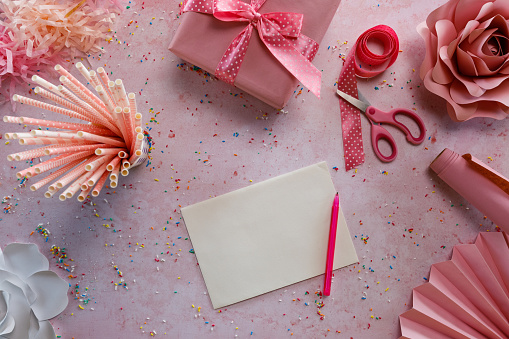 Directly above shot of a pink background with colorful sprinkles all around and a blank birthday card in the middle, surrounded with drinking straws, paper flower decorations, ribbons, a gift box and pom-poms, all pink colored.