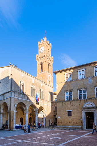 The Town Hall of Pienza is hosted in a 15th-century palace with a high civic tower, overlooking the central Piazza Pio II. Tourists strolling.