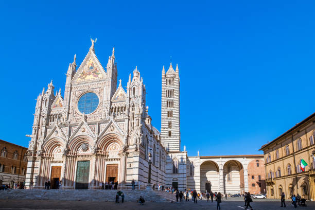 Tourists strolling in Piazza del Duomo in Siena, where overlooks the majestic Cathedral, begun in the 12th century, a masterpiece of Italian Romanesque–Gothic architecture