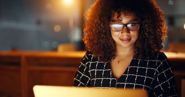Black business woman using laptop, working late in modern office with copyspace. Smiling African American female working overtime, searching for new project. Lawyer using internet research or study stock photo