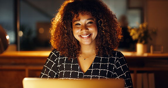 Portrait of an African businesswoman using a laptop late at night. Smiling Afro american professional working overtime on a digital campaign project, hopeful about reaching her goal or deadline