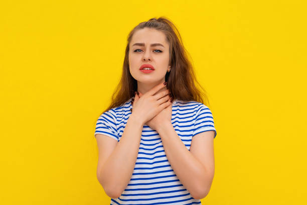 halsschmerzen. junge kranke frau, die ihre entzündete kehle hält und in weiß-blau gestreiftem t-shirt über gelbem hintergrund steht. gesundheitliche probleme - throat exam stock-fotos und bilder