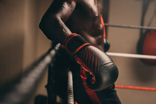 Muscular African boxer resting against the ropes of a boxing ring in a gym. Sweaty young man taking a break from training in a fitness gym.