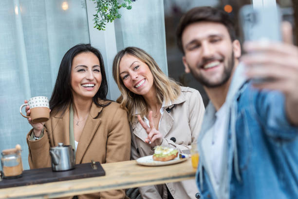 grupo de amigos geniales compartiendo un brunch juntos mientras se toman una selfie con el teléfono inteligente en la saludable terraza de la cafetería. - mobile phone coffee shop group of people women fotografías e imágenes de stock