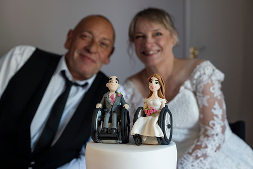 A bride and groom, who are wheelchair users that have just got married, at their evening reception in a wedding venue in Morpeth, North East England. They are out of focus, looking at the camera while smiling behind their wedding cake. The cake is in focus and has figurines of the happy couple made out of icing on top of it.