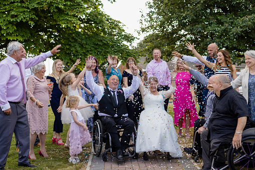 A bride and groom, who are wheelchair users that have just been married at a venue in Morpeth, North East England. They are outside with their wedding guests and the guests are throwing confetti over them while they all have their arms in the air.