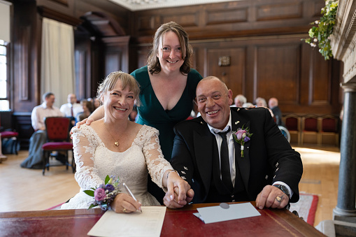 A bride and groom, who are wheelchair users, sitting at a desk in the registry office where they have just got married in Morpeth, Northumberland. They are signing the register while looking at the camera and smiling. The bride's daughter has her arm around her mum and father-in-law and is also looking at the camera and smiling.