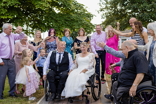 A bride and groom, who are wheelchair users that have just been married at a venue in Morpeth, North East England. They are outside with their wedding guests and the guests are throwing confetti over the newly married couple, to celebrate with them.
