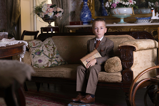 Little boy wearing a three piece suit and reading in a room full of Victorian furniture.