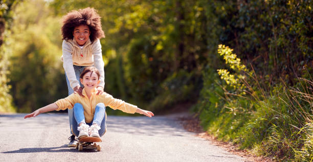 children having fun with boy pushing girl on skateboard on country road - wide screen imagens e fotografias de stock