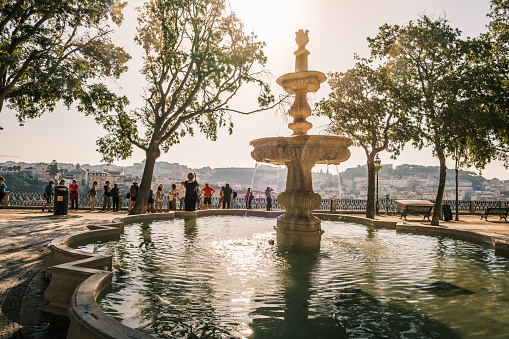 Lisboa , Portugal; 18 July 2022: Fountain in São Pedro de Alcântara park in Lisbon with a group of foreign tourists in the background contemplating the viewpoint