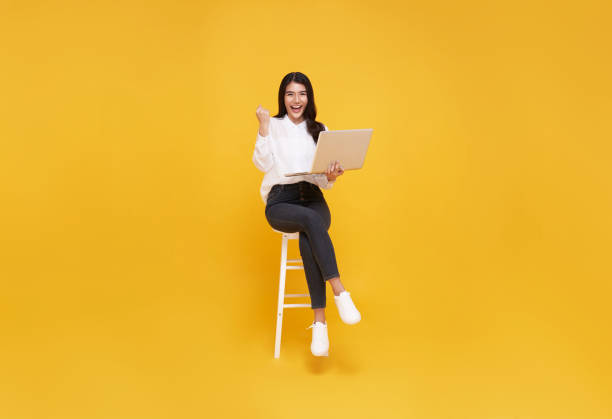 Young woman asian happy smiling. While her using laptop sitting on white chair and looking isolate on yellow background. stock photo