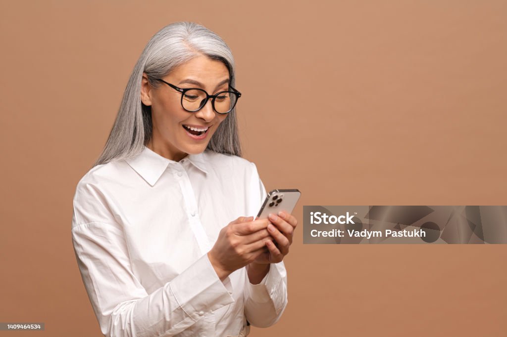 Surprised attractive senior woman holding smartphone, chatting in social networks, texting messages, smiling, reading good news Surprised attractive senior woman holding smartphone, chatting in social networks, texting messages, smiling, reading good news, isolated on beige Surprise Stock Photo