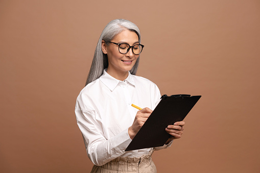 Senior confident business woman writing at the clipboard. Mature female employee in formal wear isolated on beige