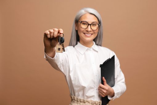 Happy senior asian business woman professional Real Estate Agent real estate agent wear suit looking at camera, holding key to new property house and clipboard isolated on beige background copy space