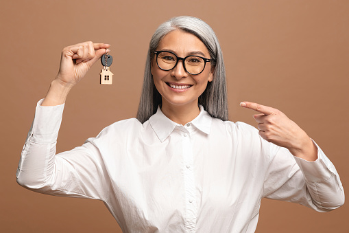 Happy overjoyed senior woman holding keys with keychain in form of little house isolated on brown background. Smiling woman holding keys from new property, happy buyer of own estate. Lady relocated