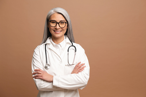 Smiling gray-haired Asian female doctor pediatric, physical, therapist wearing white medical gown with stethoscope stands with arms crossed isolated on brown, copy space. Healthcare and medicine