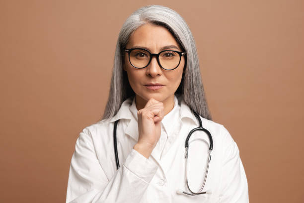 doctora asiática seria de mediana edad con un estetoscopio en un abrigo médico posando aislado sobre marrón - doctor thinking asking pensive fotografías e imágenes de stock