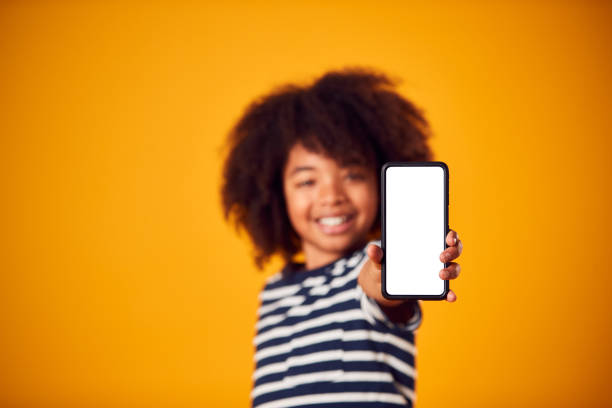 portrait en studio d’un garçon montrant un téléphone portable avec un écran de médias sociaux sur fond jaune - elementary age focus on foreground indoors studio shot photos et images de collection